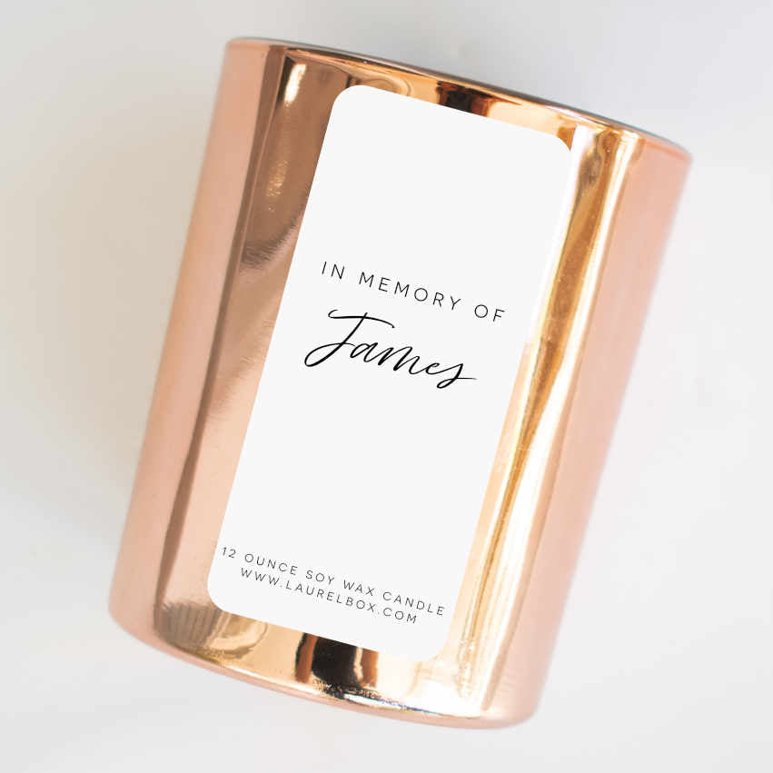 A gold-coated candle with a white sticker with the text In Memory of James laying flat tilted to the right on a white surface