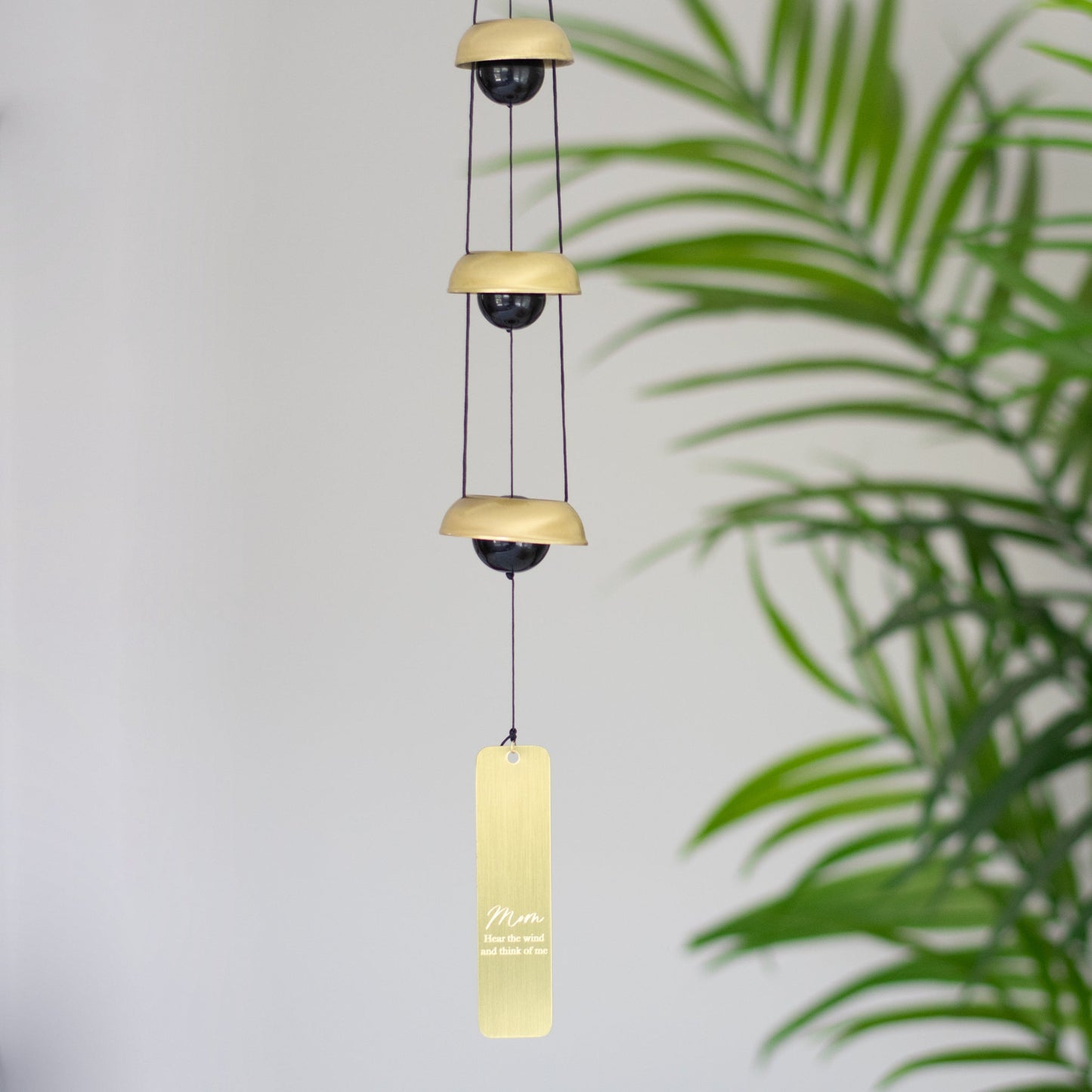 A wind chime hanging inside next to a plant that features three metallic bells, black cords, and a vertical rectangular tag at the bottom with the text mom and Hear the wind and think of me